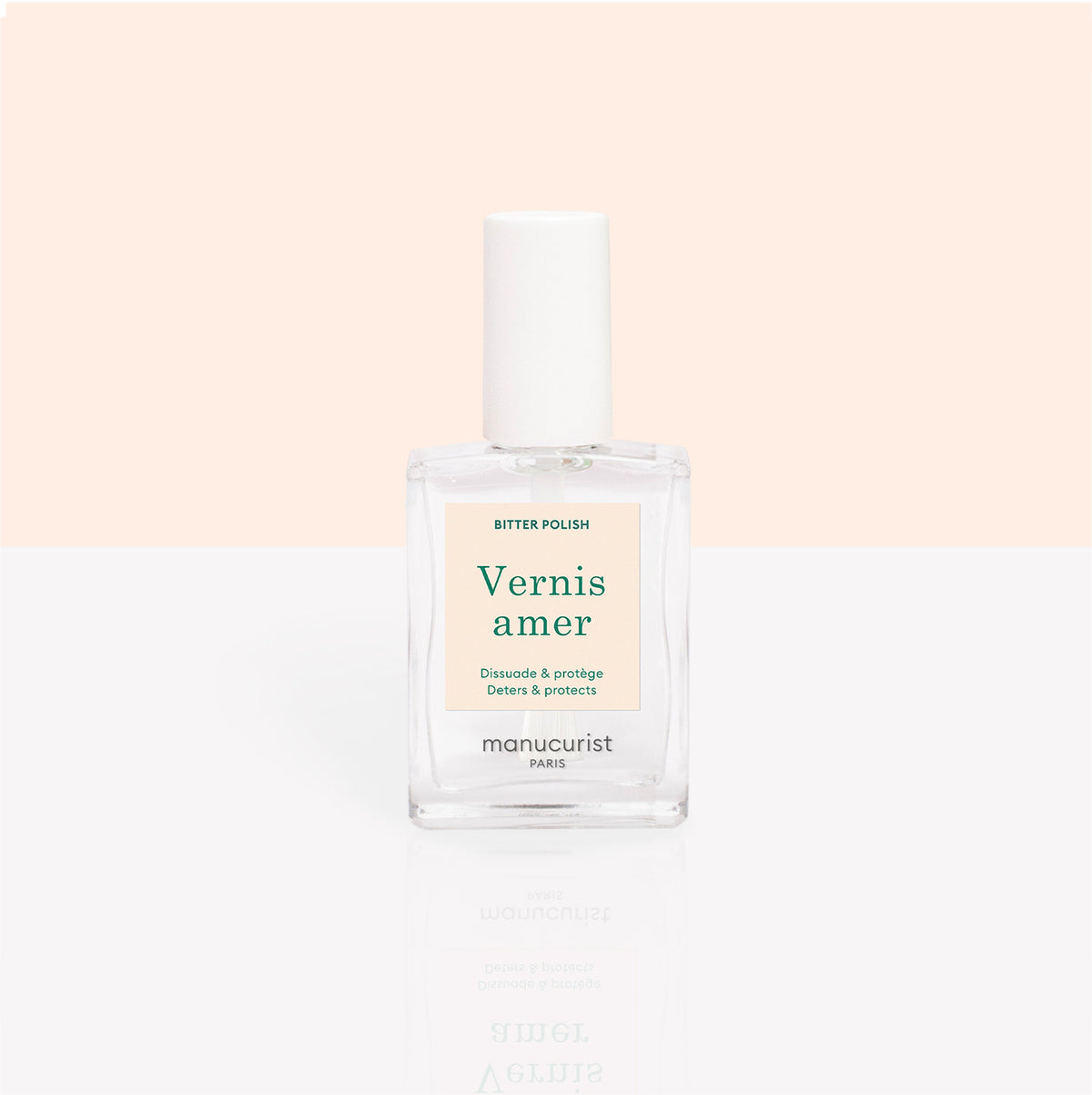 A clear glass bottle of Manucurist Bitter Polish, an anti-nail-biting solution with a white cap, is shown against a pastel peach and white background. The label features green and gold text.