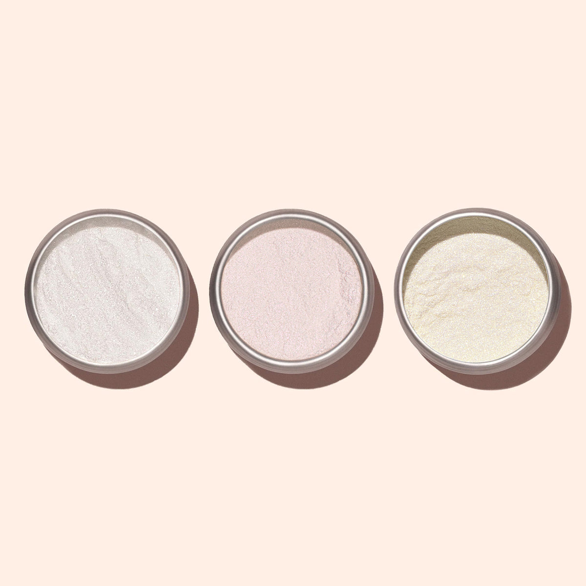 Three round metal bowls are arranged in a row, each with a different fine powder—white, light pink, and pale yellow Manucurist Golden Glazed Powder—displaying pearly gold shimmer on a light beige background.