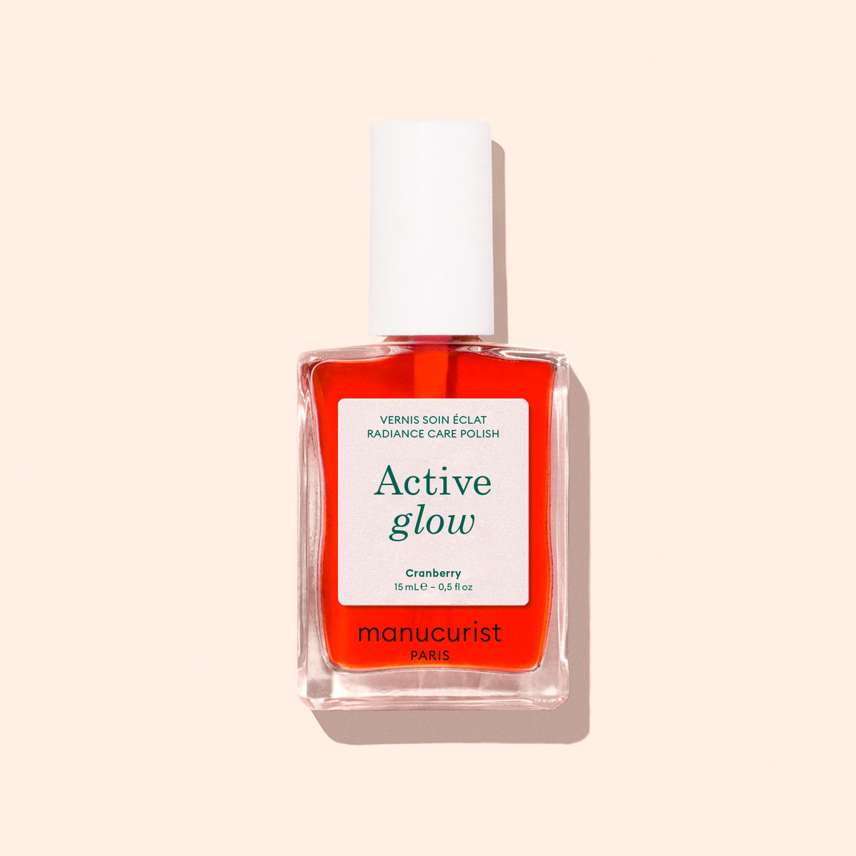 A clear bottle of Manucurist Active™ Glow Cranberry nail treatment, featuring a plant-based formula and a white cap, is set against a light beige background.