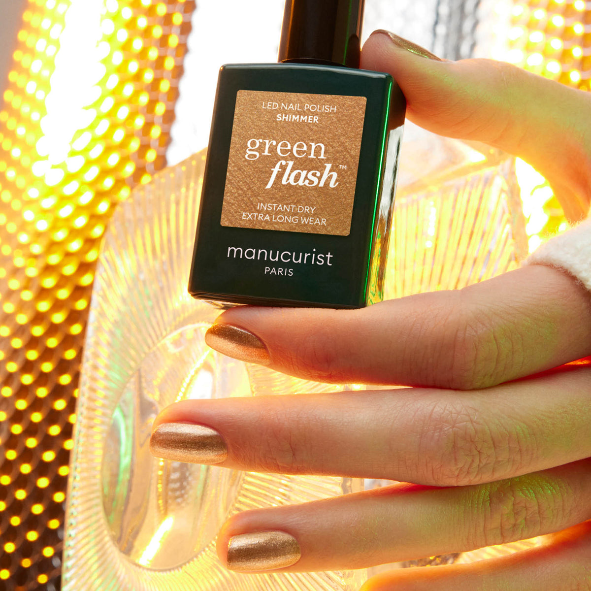 A hand with honey-bronze gel polish holds a bottle of Manucurist Shimmer LED nail polish. The background glows warmly, accented by a decorative glass piece.