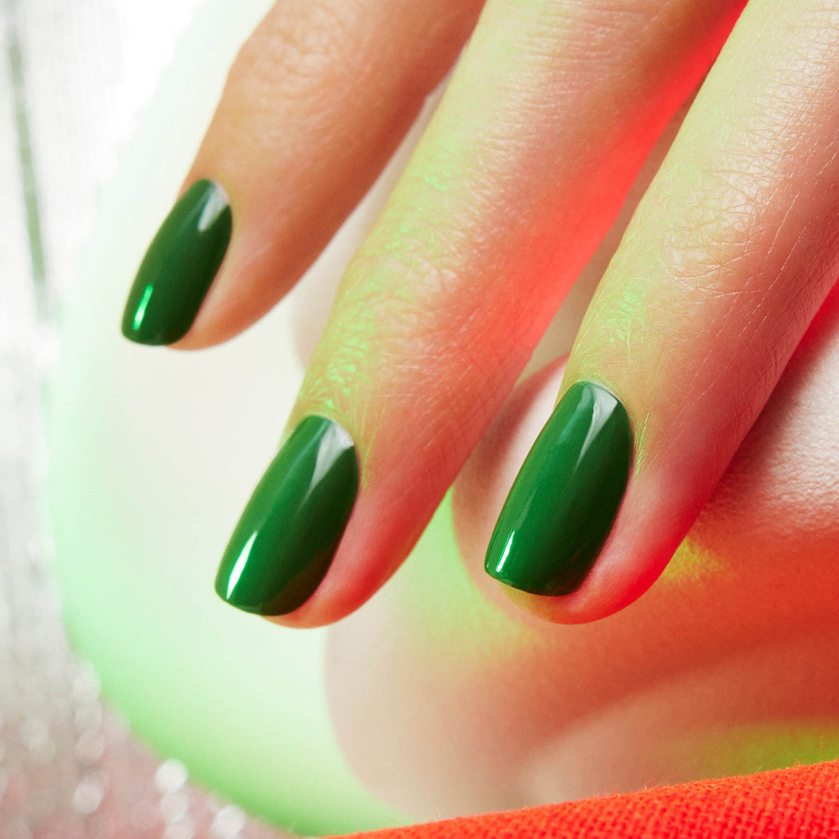 A close-up of a neatly manicured hand showcases glossy, cool-toned mineral green "Jade" nail polish by Manucurist against a blurred background with red and green lighting.