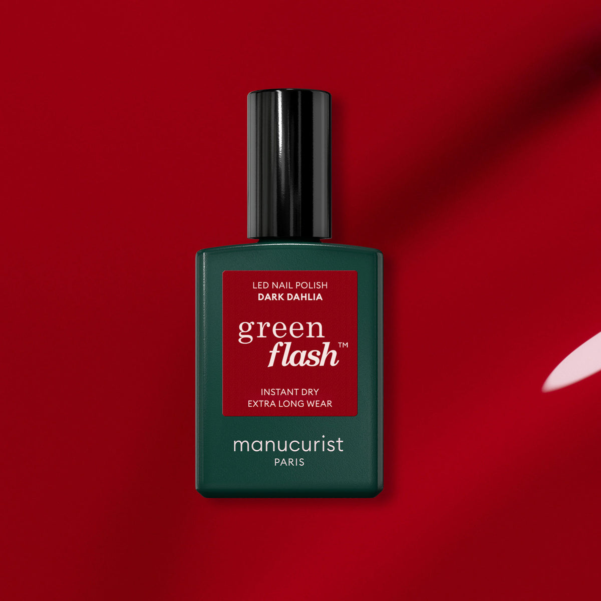 A bottle of Manucurist Dark Dahlia gel polish sits on a rich red background, displaying its dark green body, black cap, and red label.