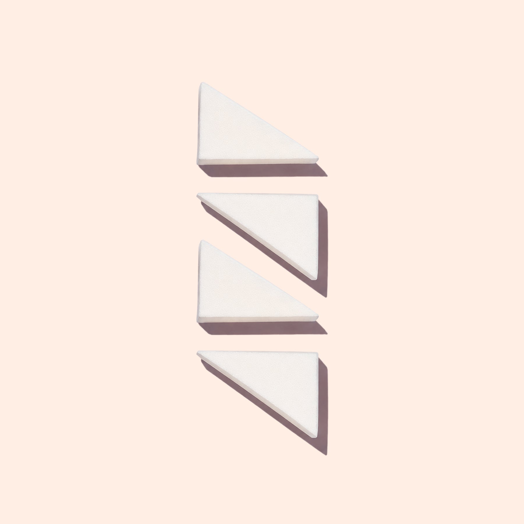 Three white triangular wedges from Manucurist’s Nail Art Sponges - x4 are arranged vertically on a light beige background, each casting a shadow to the lower right.