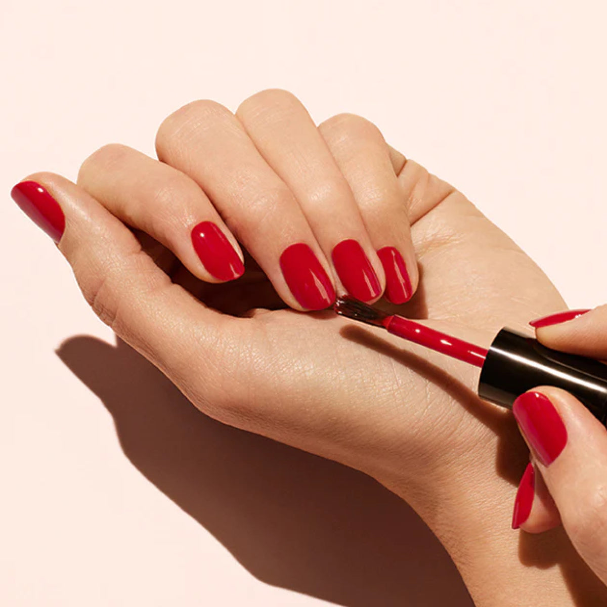 A person is painting the nails on their left hand with bright red polish from the Manucurist Basic Kit, using a brush applicator in their right hand. The background is light and neutral.
