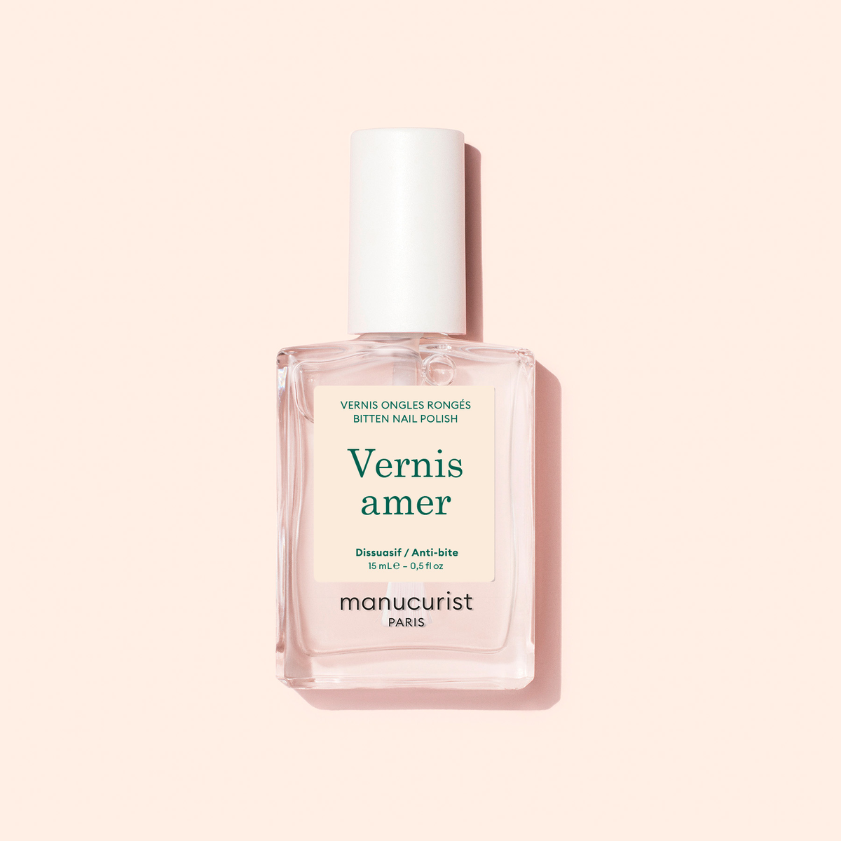 A bottle of Manucurist Bitter Polish with a white cap, labeled “Vernis amer, Dissuade protège, Deters protects,” rests on a pale peach background—offering an effective anti-nail biting solution.