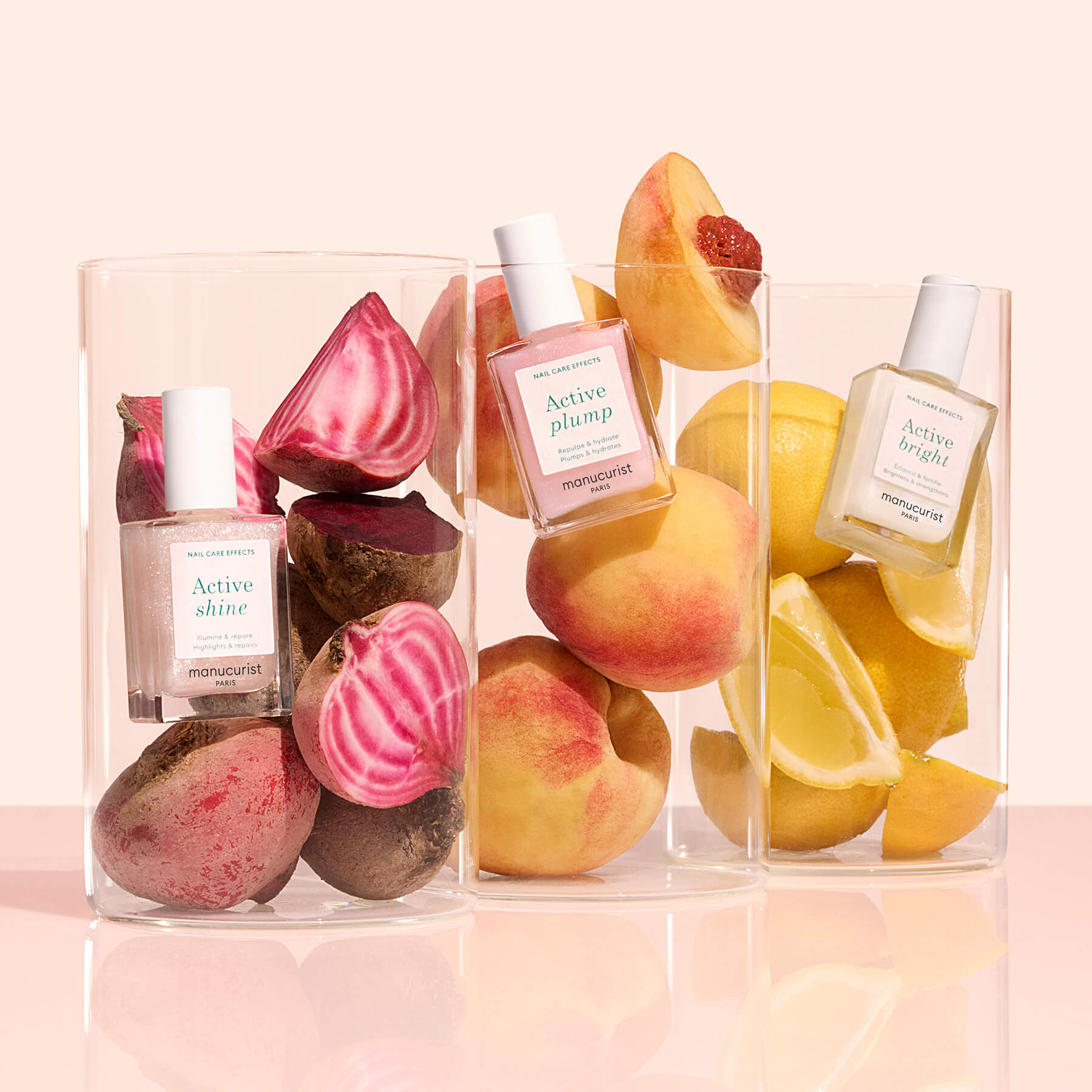 Three glass containers of Manucurist’s Customisable Rainbow Active™ polishes are shown beside beets, peaches, and lemons on a light pink background for a personalised look.