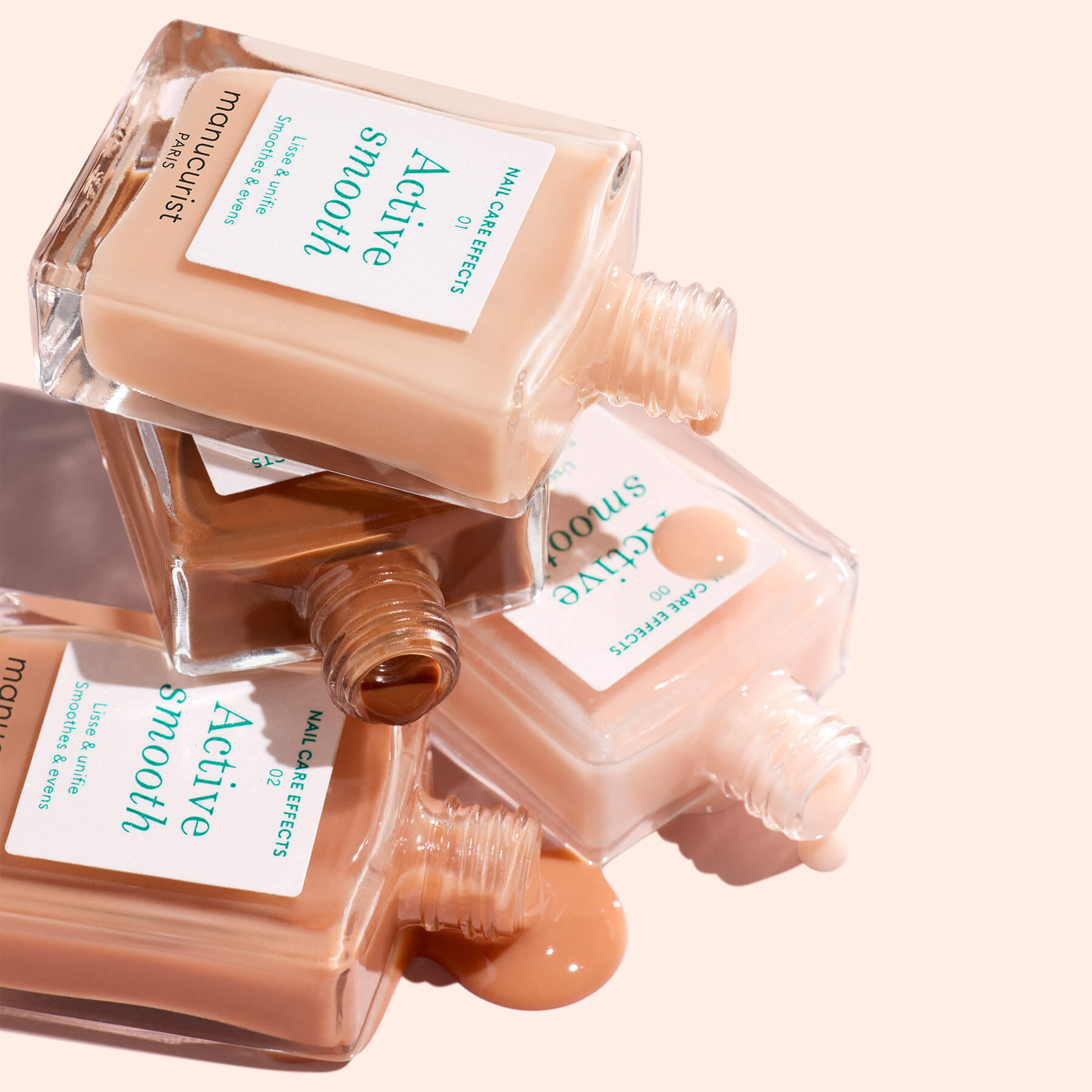 Three bottles of Manucurist Customisable Rainbow Active™ nail polishes lie on their sides, caps off, spilling light beige and tan polish onto a light peach surface for a personalised touch.