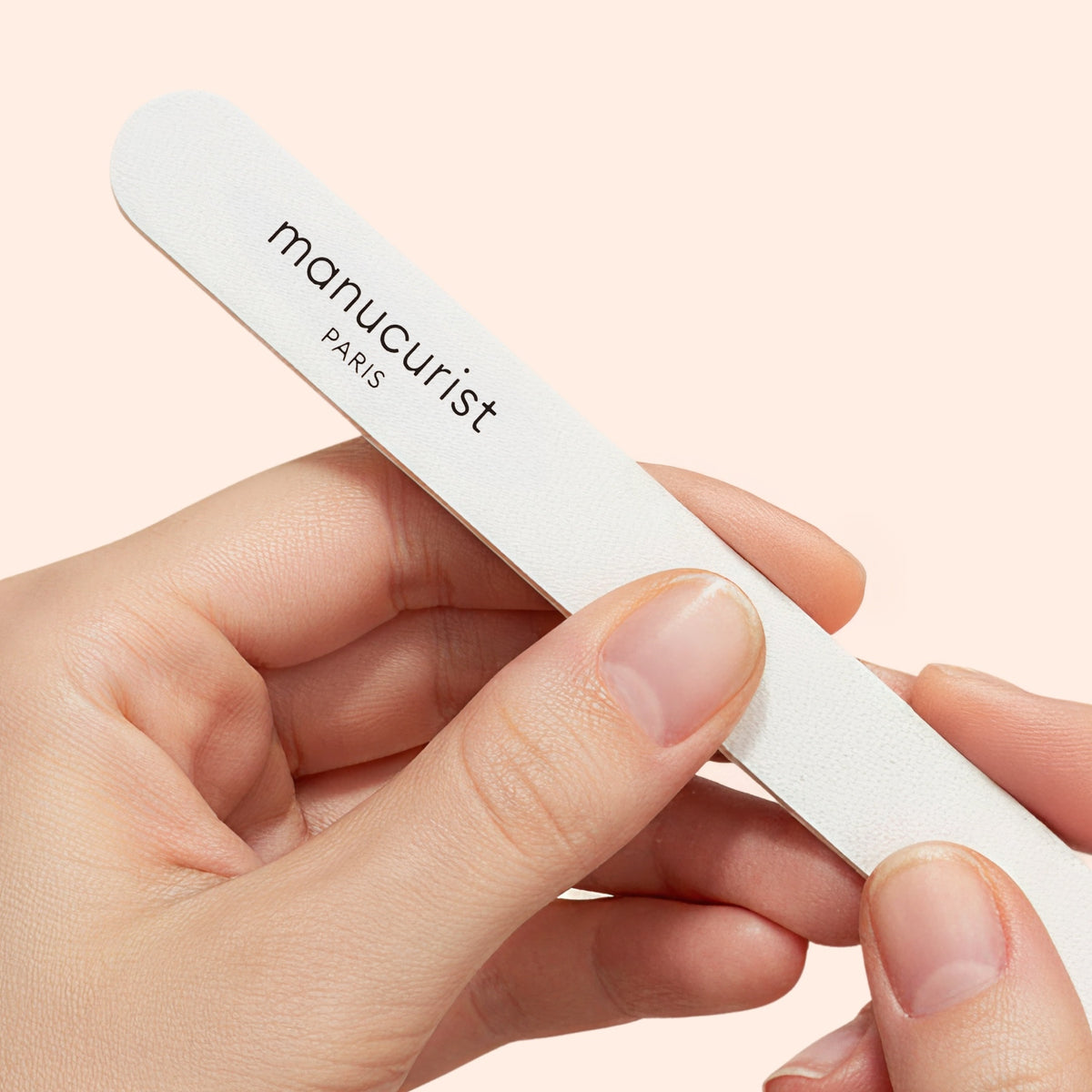 A close-up of hands holding a Manucurist 180/240 emery board for fragile nails (set of 5), ideal for shaping sensitive or brittle nails, with the brand name printed on it against a light beige background.