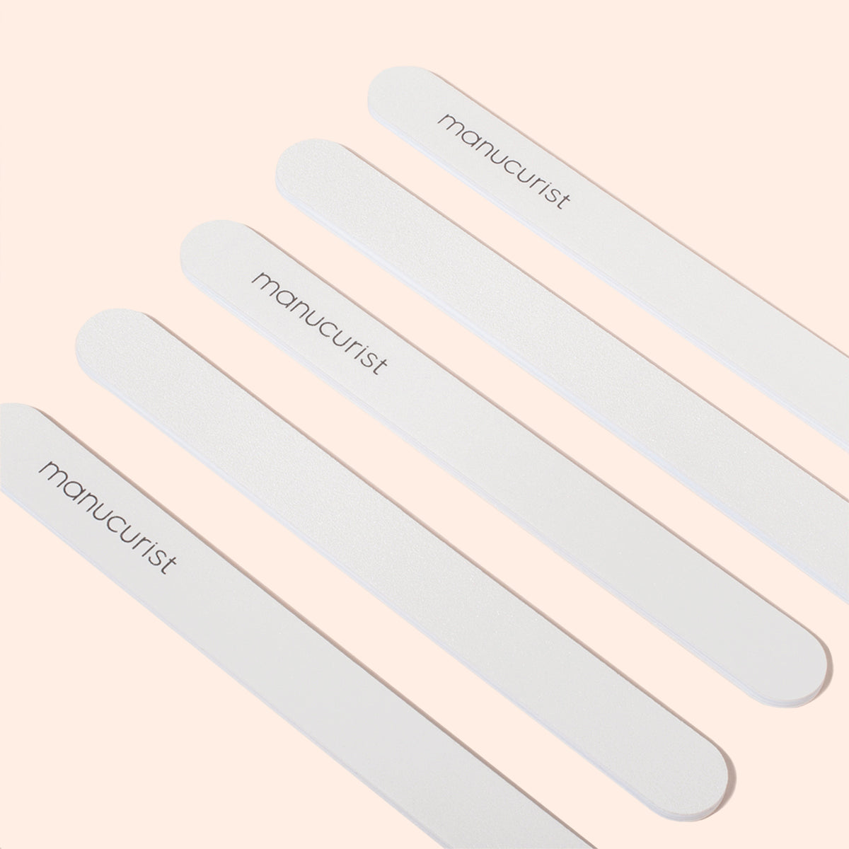 Five white emery boards with rounded ends, each labeled Manucurist, are arranged diagonally on a pale peach background—perfect for gently smoothing delicate nails.