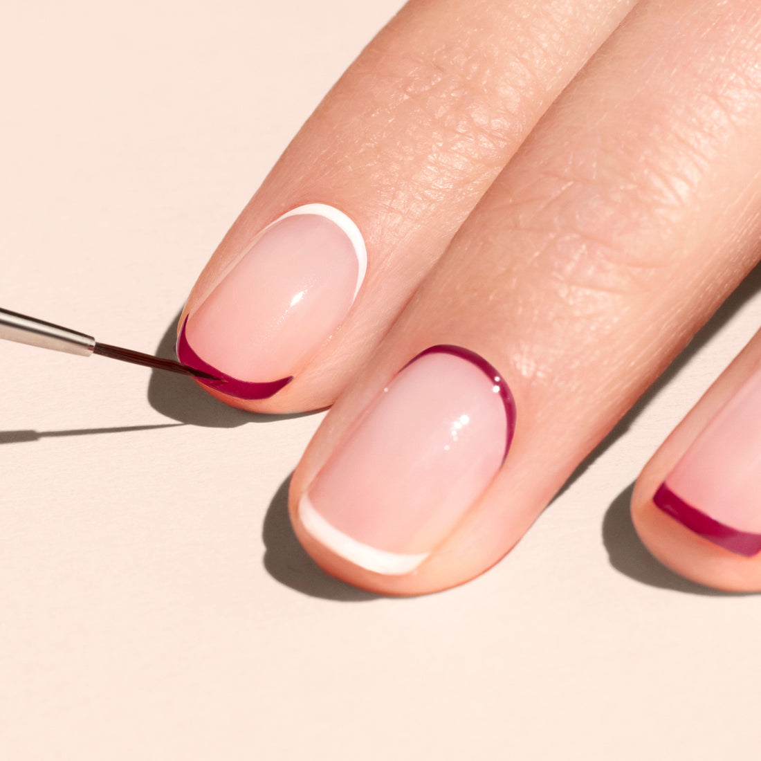 A hand with neatly manicured nails features minimalist nail art with thin white and dark red lines. The Manucurist Liner Brush is used to apply dark red polish to one nail near the tip and cuticle.