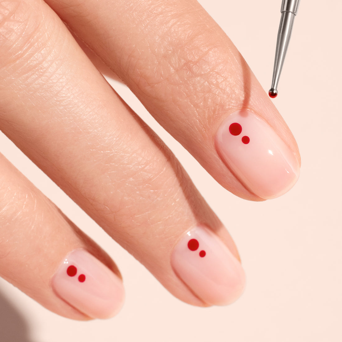A hand with light pink polished nails, each adorned with small red dots, is shown as the Manucurist Dotting Tool precisely applies a red dot to one nail. The background features a light, neutral shade.