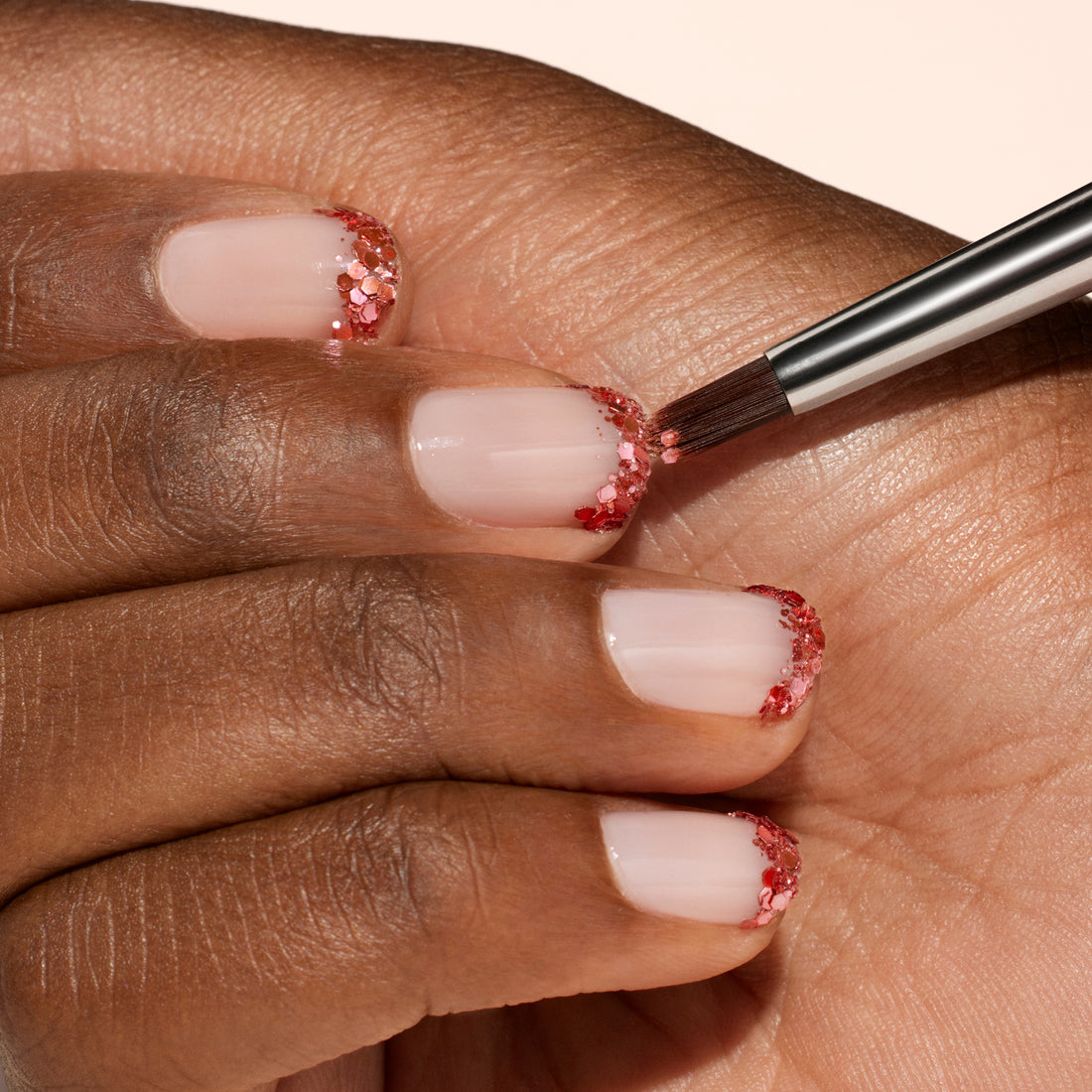A close-up of a hand with nude nails and red glitter tips. The Manucurist Glitter Brush, with vegan synthetic bristles, is used to apply glitter to one nail, showcasing a radiant manicure in progress.