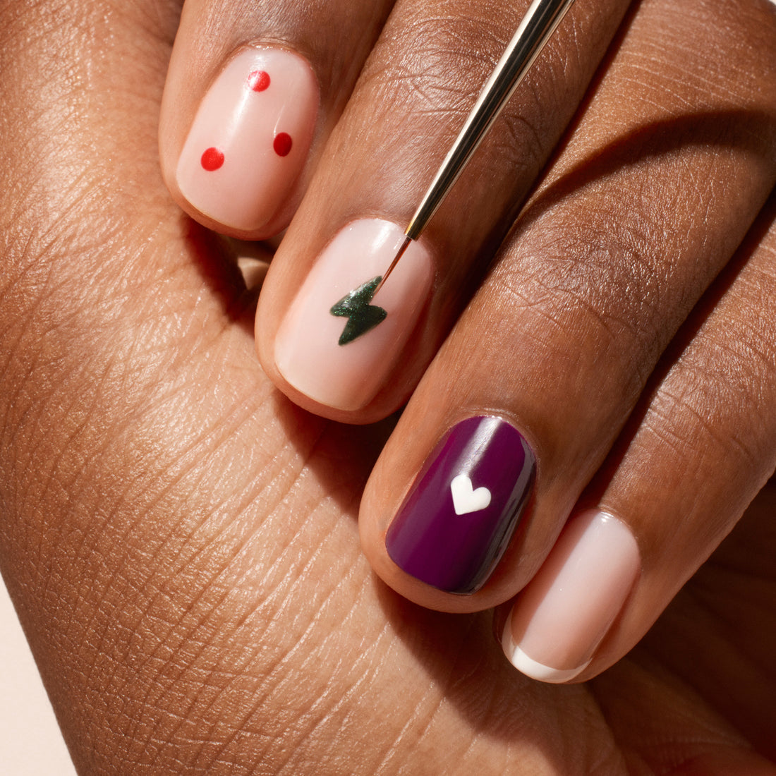 A hand with manicured nails features creative nail art—red dots, a green lightning bolt painted using the Manucurist Fine Tip Brush, and a purple heart—while the other nails are left natural with clear polish.
