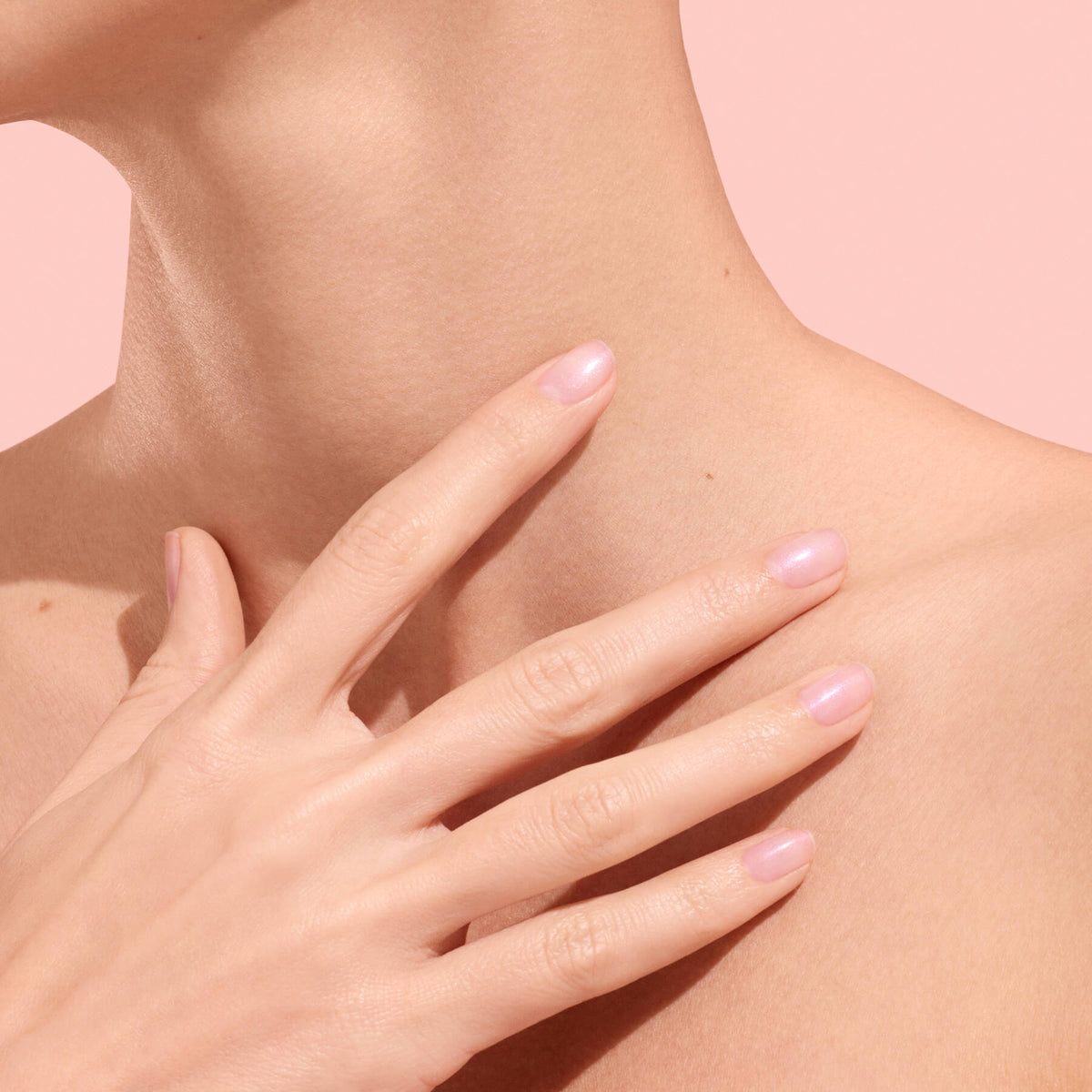 A close-up of a person’s neck and collarbone area with their hand—showcasing beautifully hydrated nails thanks to Manucurist’s Active™ Plump Pink Glazed—gently touching the skin, set against a soft pink background.