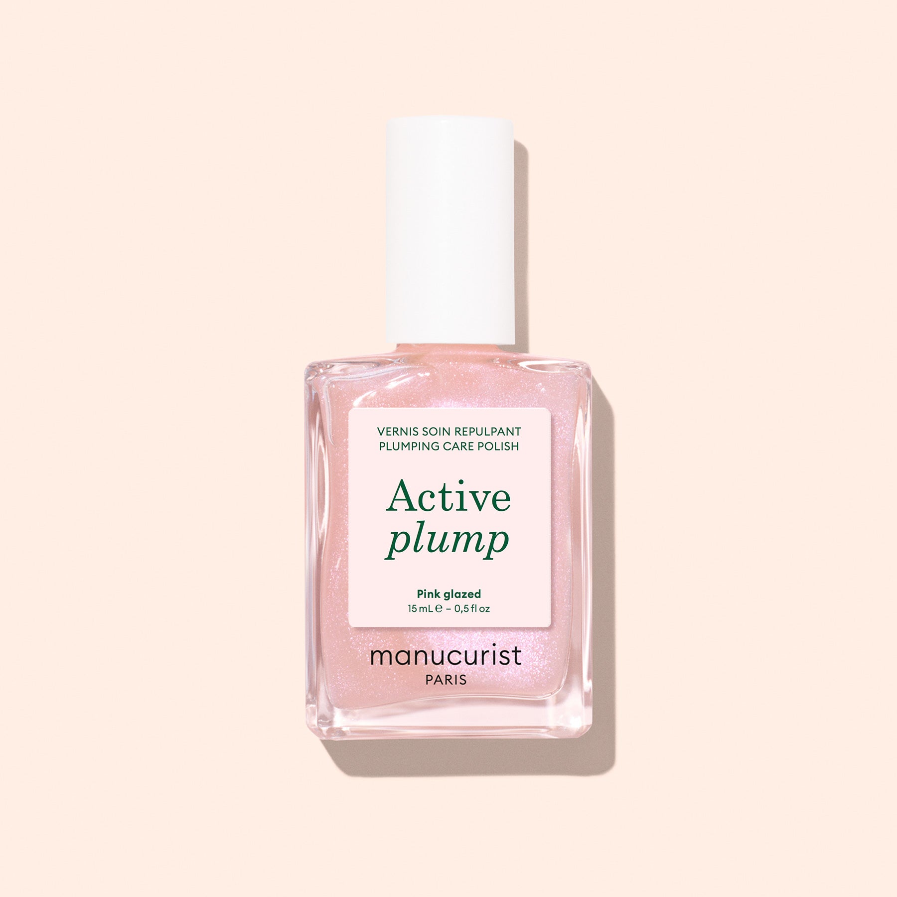 A bottle of Manucurist Active Plump Pink Glazed nail polish with a white cap and light pink formula sits on a pale peach background, its label showcasing plumping and hydrating benefits for beautifully moisturized nails.