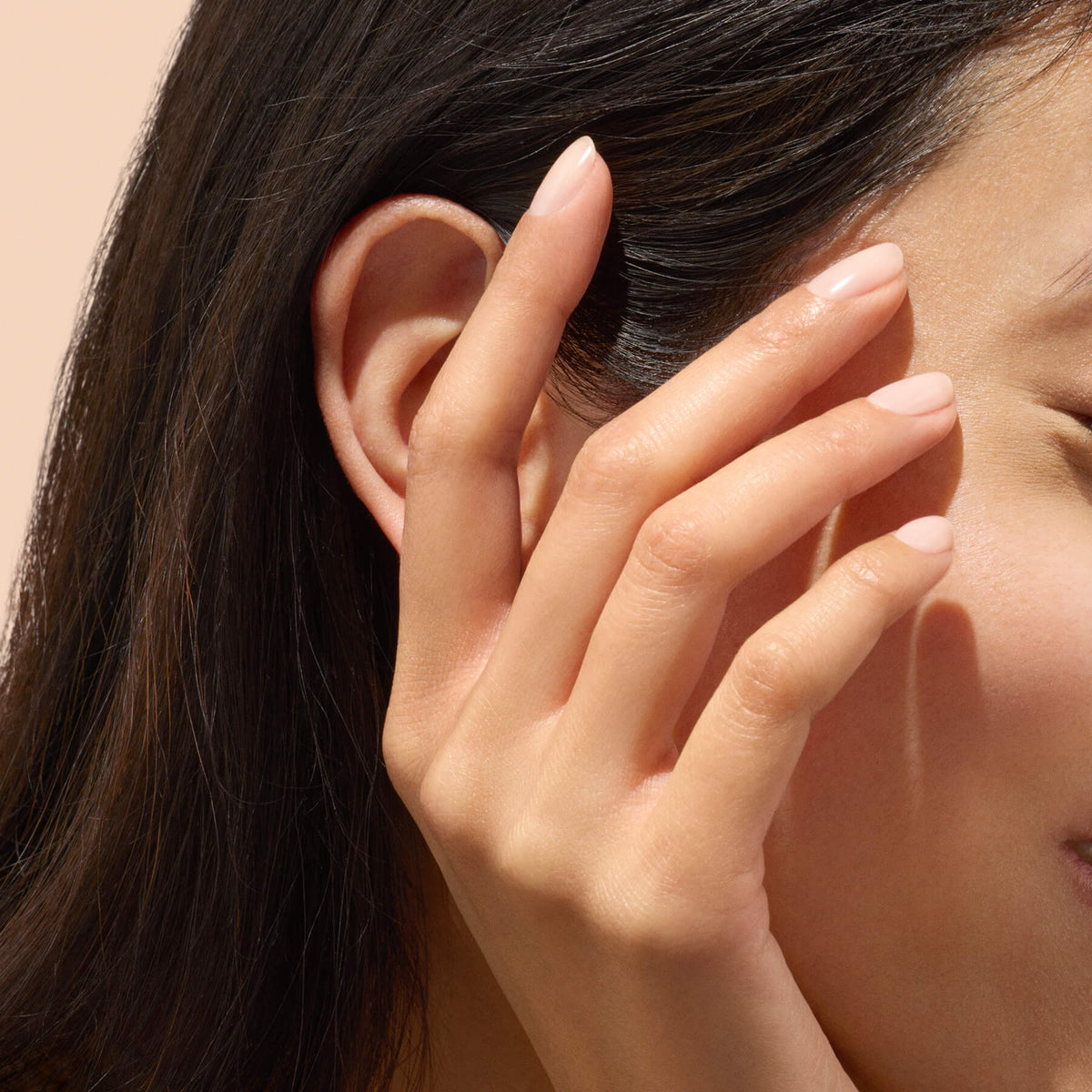 A close-up of a woman’s ear and hand with natural, nude-colored nails enhanced by Manucurist’s Active™ Smooth 01. Her fingers—reflecting thoughtful, plant-based nail care—gently touch her hair, and her skin appears smooth and healthy against a soft beige background.