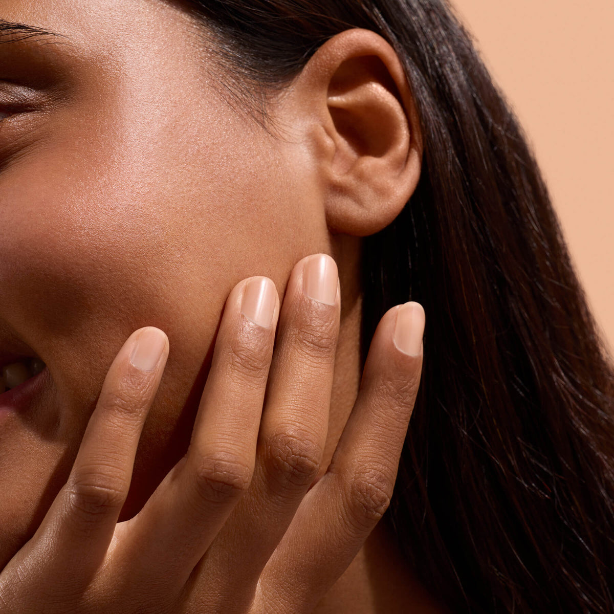 A close-up of a smiling person with smooth skin gently touching their cheek with neatly manicured fingers, highlighting the effects of Manucurist Active™ Smooth 02 nail care. The background is a soft beige color.