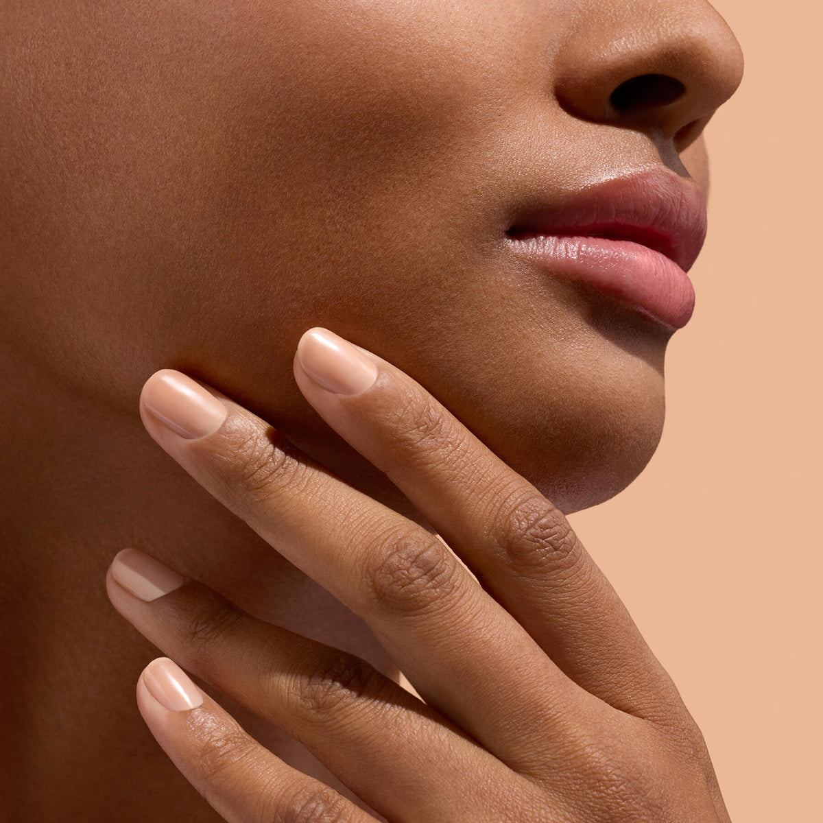 A close-up of a woman’s lower face and hand, featuring smooth skin and natural makeup, showcases her well-manicured nails treated with Manucurist Active™ Smooth 02, highlighting her commitment to nail care against a beige background.