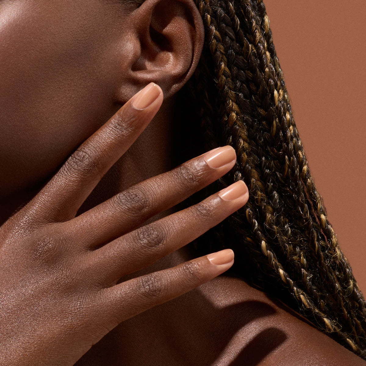 A close-up of a person’s hand, featuring neatly manicured nails painted with Manucurist’s Active™ Smooth 03—a nude shade with a subtle bronze satin finish—rests on their face. The individual has brown skin and braided hair with golden highlights, set against a warm brown background.