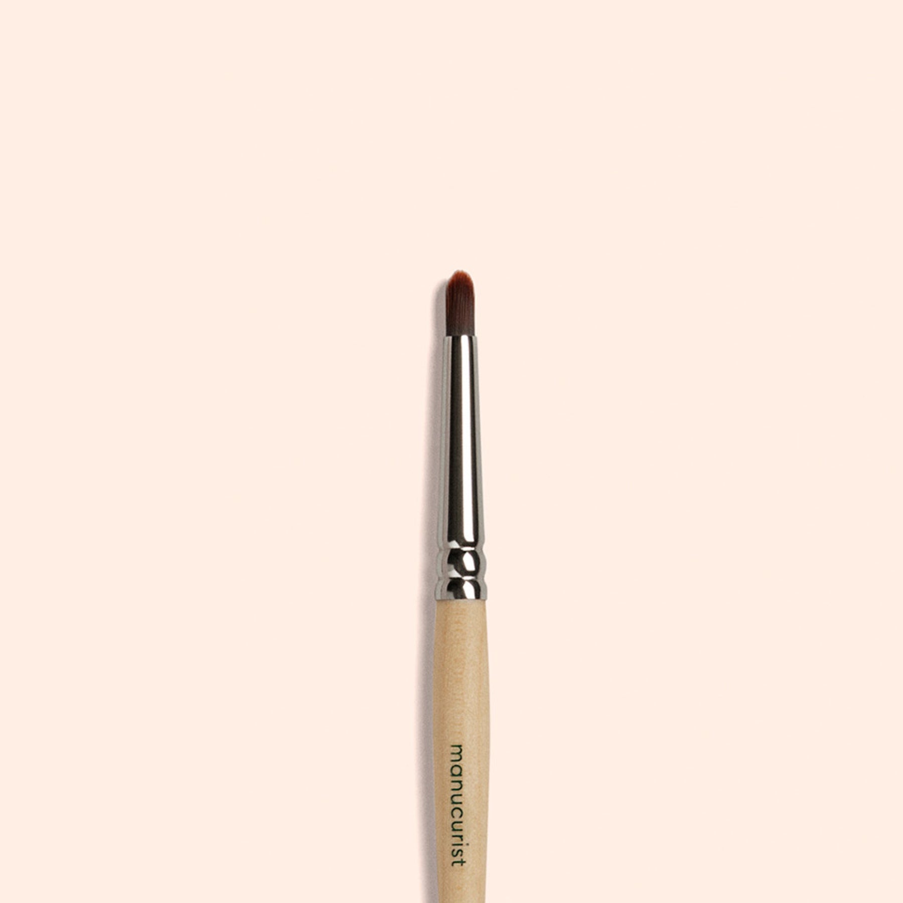 The Manucurist Glitter Brush, featuring a wooden handle, metal ferrule, and rounded vegan bristles, stands vertically on a light beige background—ideal for creating radiant nail art.