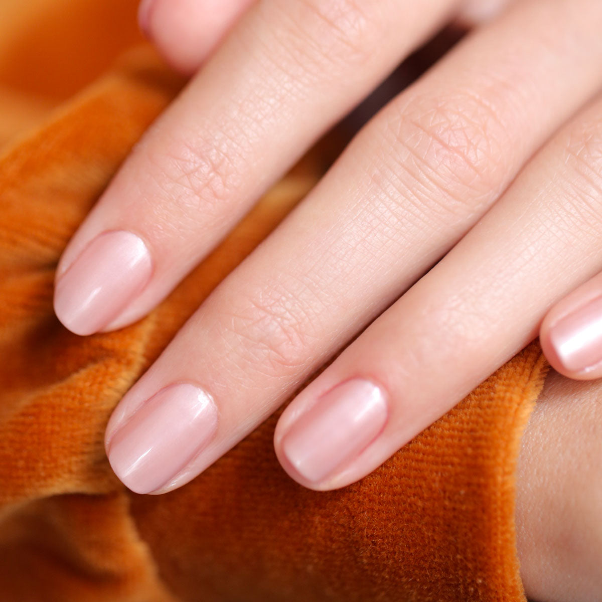 A close-up of neatly manicured nails painted in Manucurist’s Carnation, a pearly pink-beige nail polish with a shiny, natural finish, as the hand rests on soft orange fabric.