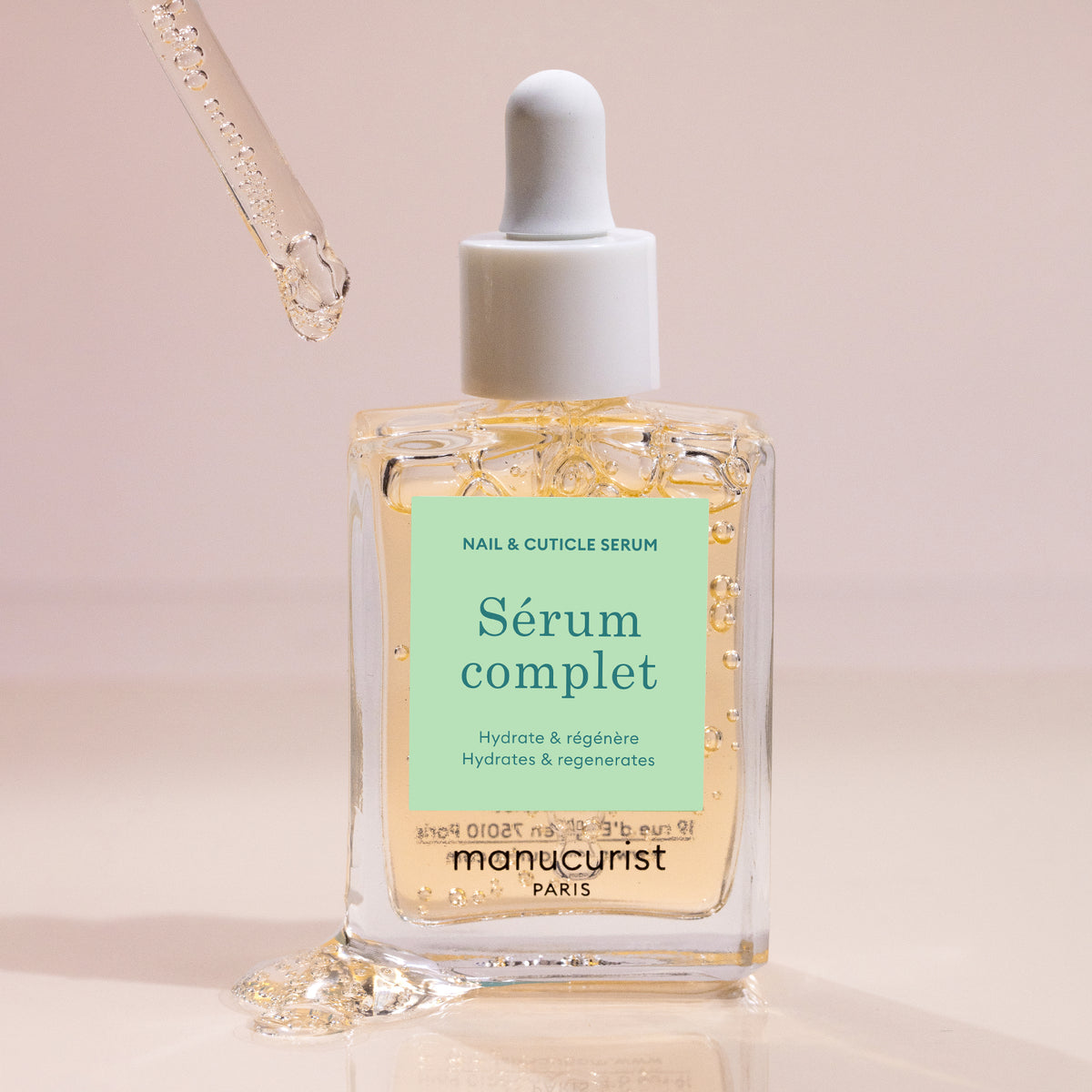 A glass dropper dispenses hydrating serum into a clear bottle labeled Complete Serum by Manucurist, a nail and cuticle treatment that hydrates and regenerates, set against a soft beige background.