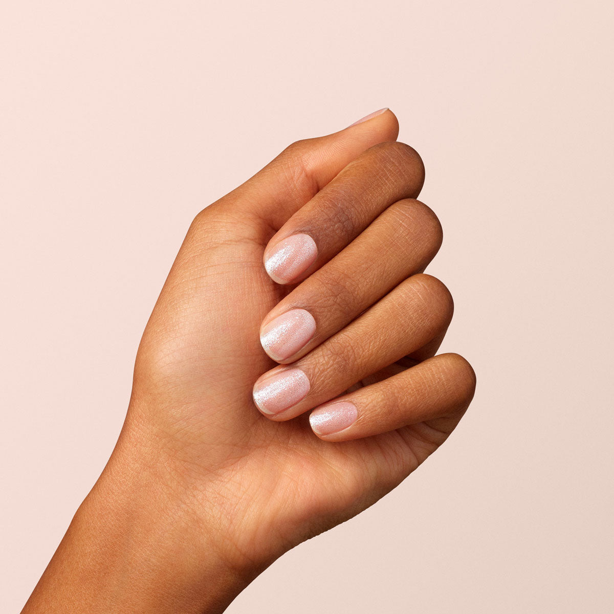 A hand with medium brown skin displays neatly manicured nails painted in Manucurist’s Diamant, a pale pink shimmery silver polish, against a plain light beige background.