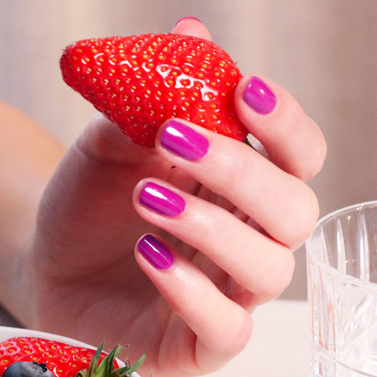 A hand with glossy, pinky mauve Armeria nail polish by Manucurist holds a ripe strawberry near a clear glass. The softly blurred background reveals another strawberry and a blueberry on the table.