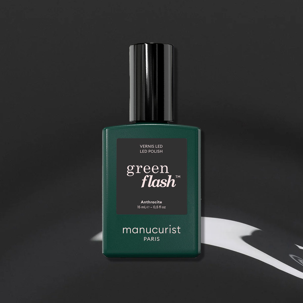 A dark green rectangular Manucurist bottle of Green Flash gel polish in the Anthracite shade, a chic dark grey, with a black cap sits on an abstract black and white background.