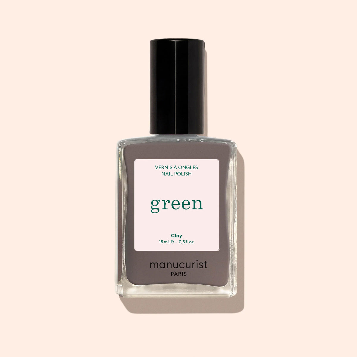 A bottle of Manucurist Clay nail polish, a chestnut gray shade from the Green™ range labeled Quick Dry and Intense Color, with a black cap, is shown against a pale beige background.