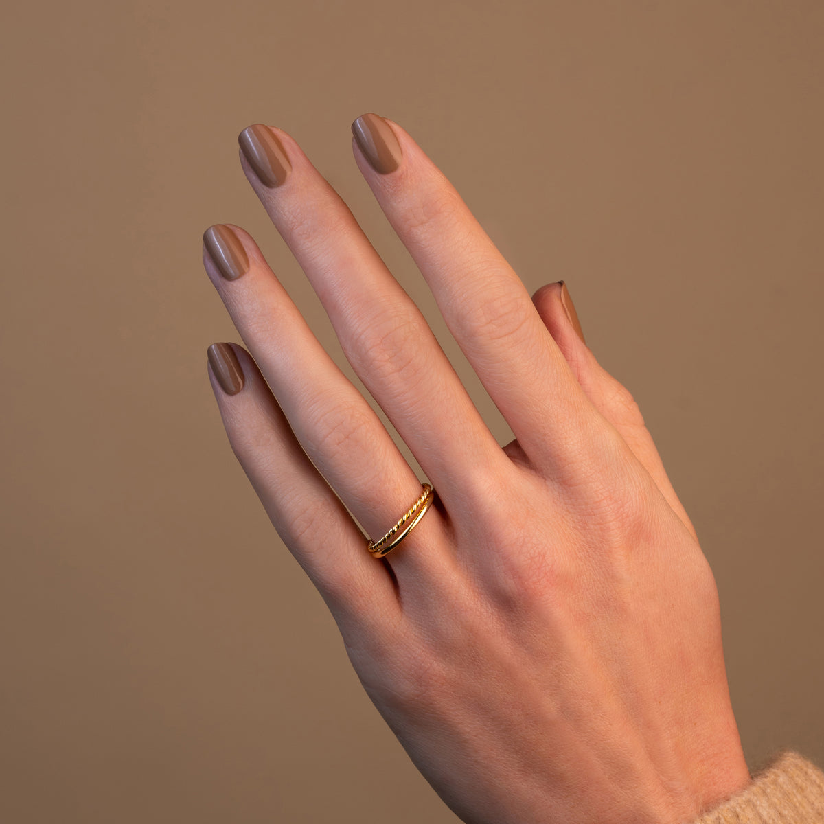 A hand with neatly manicured Orme by Manucurist café au lait nail polish wears a simple thin gold ring on the middle finger, set against a plain beige background.