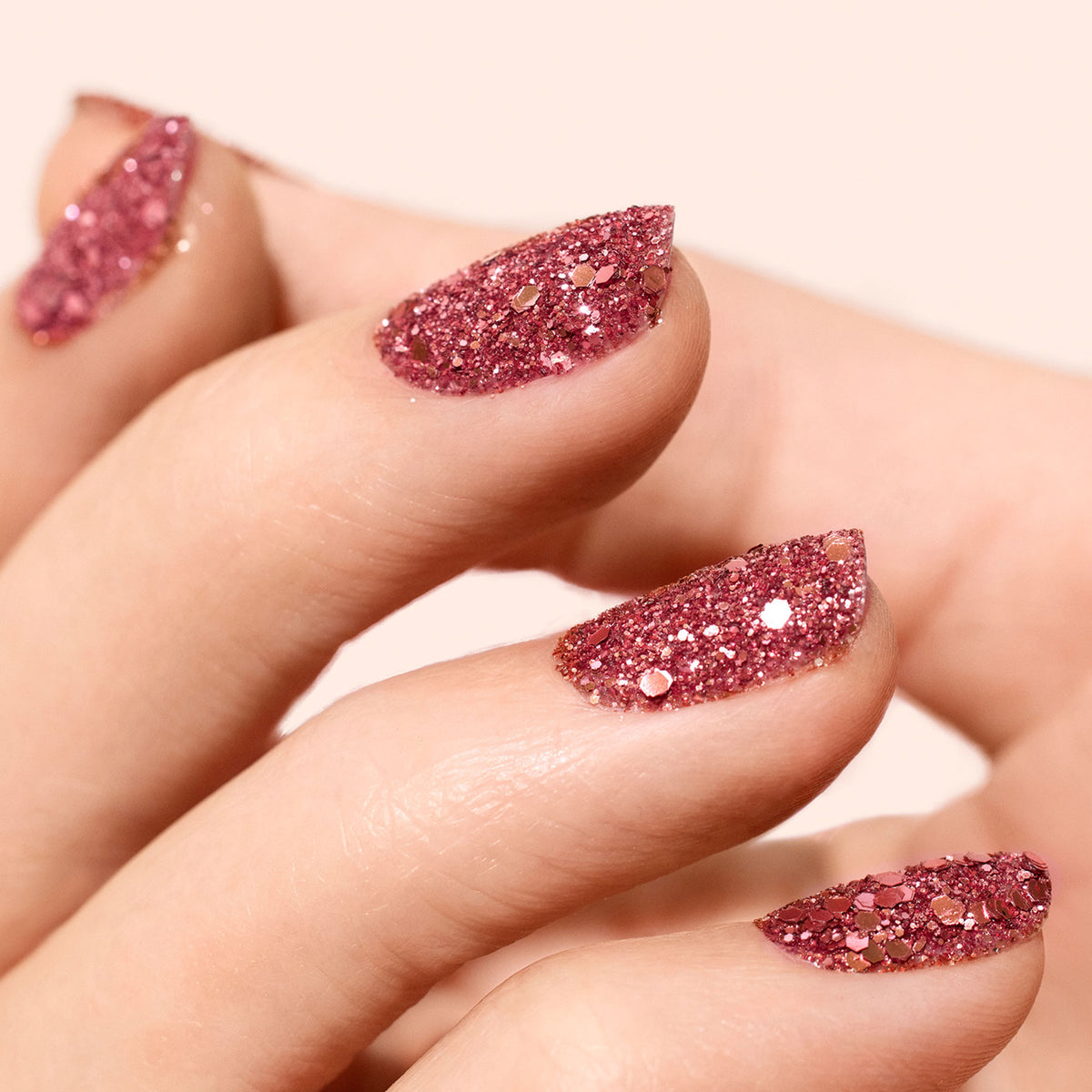 A manicured hand features almond-shaped nails painted with Manucurist Pink Quartz biodegradable glitter, creating a sparkling, shimmering effect as the light catches the eco-friendly gel polish.