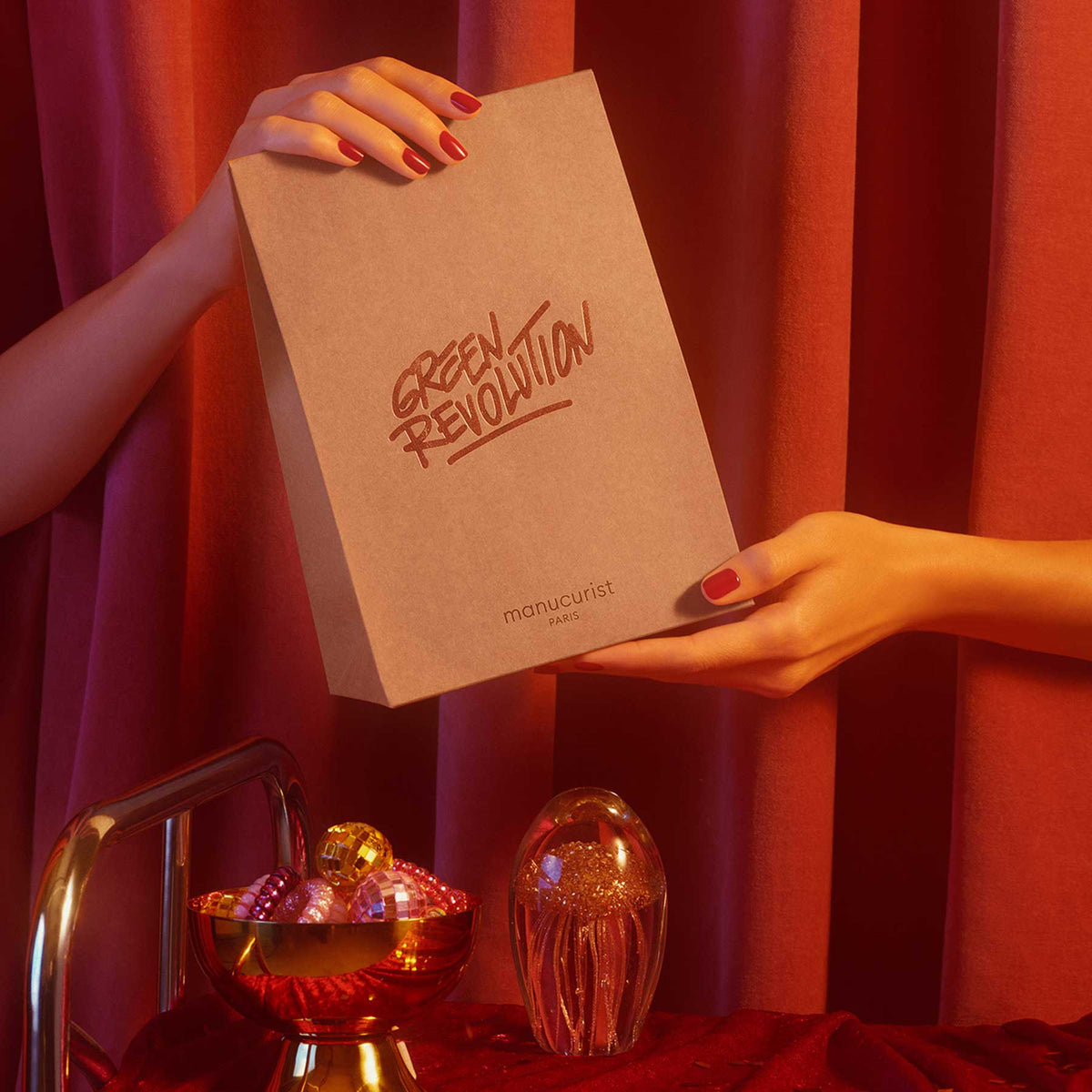 A person with red manicured nails holds a brown Manucurist box beside a gold bowl of ornaments and a Manucurist Gift bag - S, all on red velvet against deep red curtains.