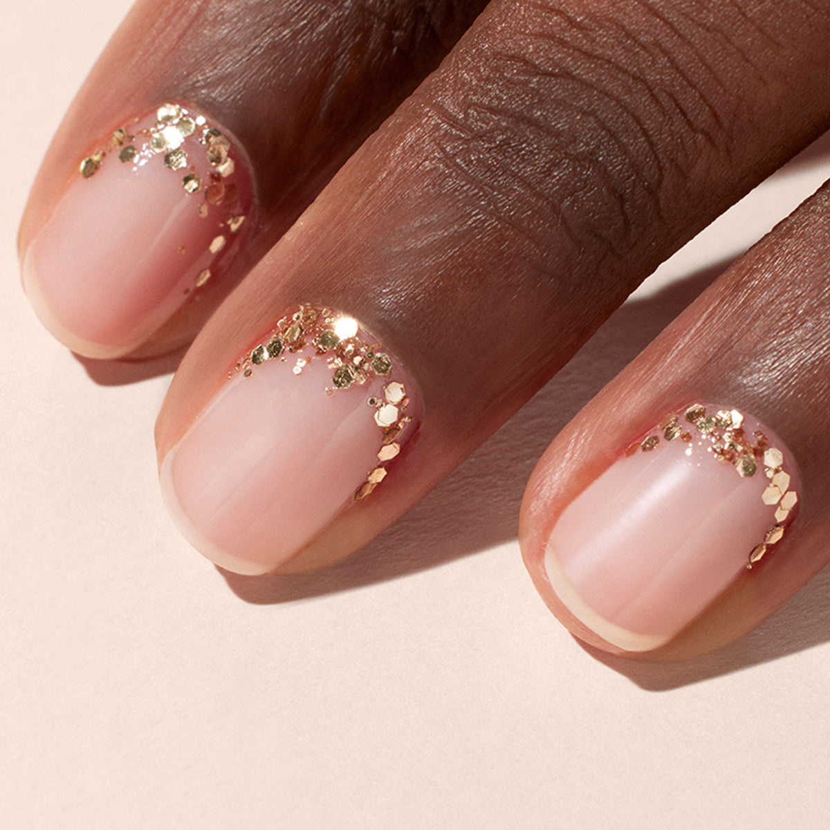 A close-up of three fingers with short, neatly filed nails featuring a nude base and Manucurist Rose Gold biodegradable glitter accents along the cuticle area.