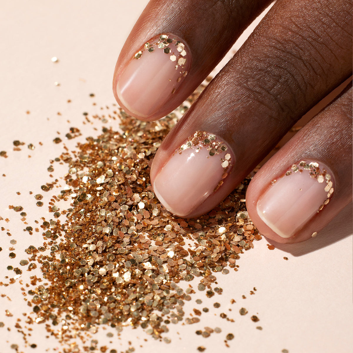A hand with brown skin shows nails painted in a nude polish and accented near the cuticles with Manucurist Rose Gold biodegradable glitter, some of which is also scattered on the pale surface beneath the fingers.