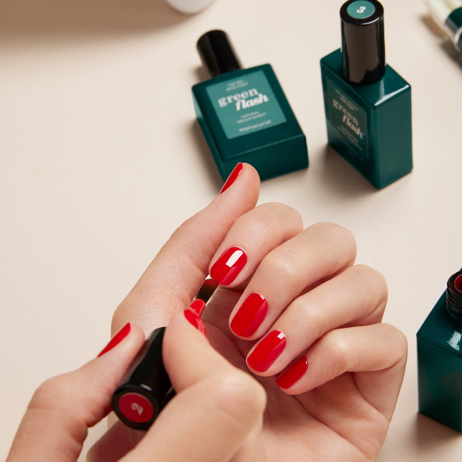 Someone applies bright red polish from the Manucurist Green Flash Set to their nails, with Green Flash gel polish bottles and manicure essentials nearby on a neutral surface for a flawless finish.