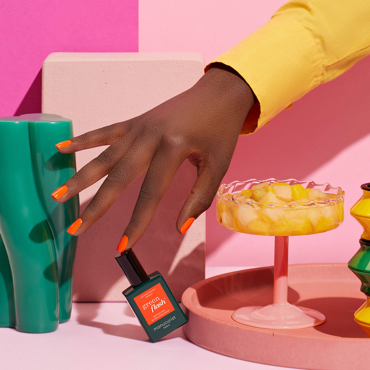 A hand with orange-painted nails holds a bottle of Manucurist Sunset nail polish. In the background are colorful objects—a green vase, a fruit dessert dish, and pink and yellow geometric shapes.