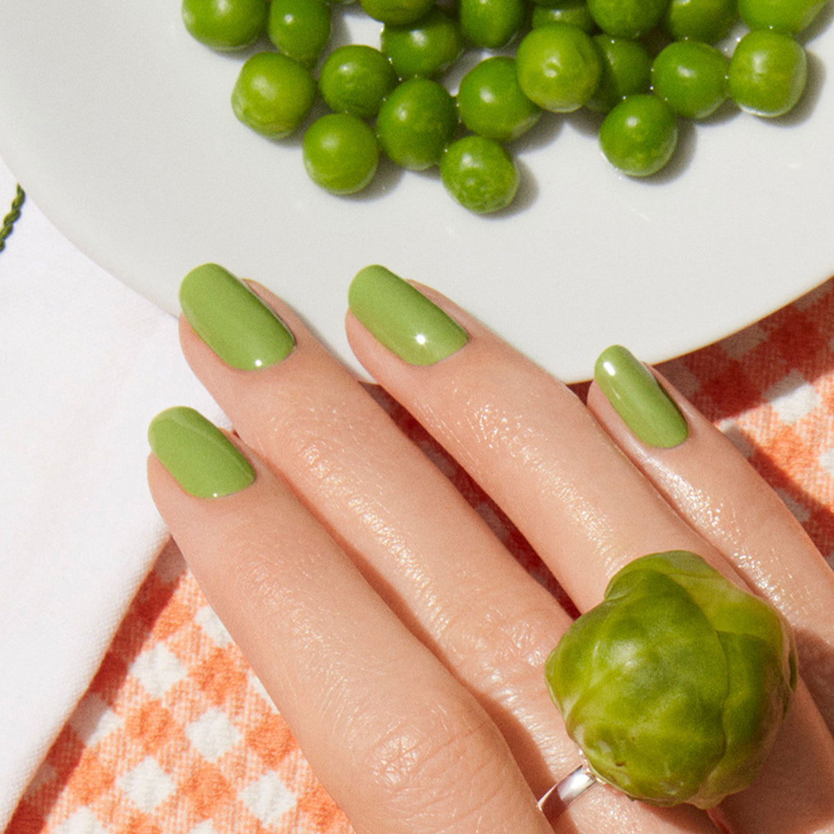 A hand adorned with Manucurist’s Sweet Pea light green nail polish and a Brussels sprout ring rests on an orange-and-white checkered cloth beside a plate of green peas.
