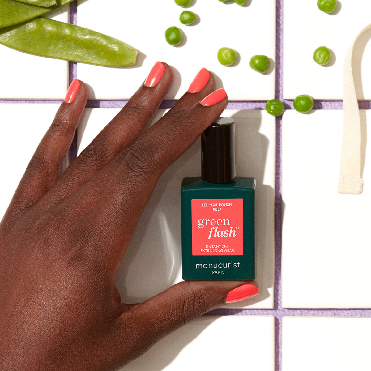 A hand with coral-colored nails holds a bottle of Manucurist Pulp gel polish. Green peas and snap pea pods are scattered on a white tiled surface.