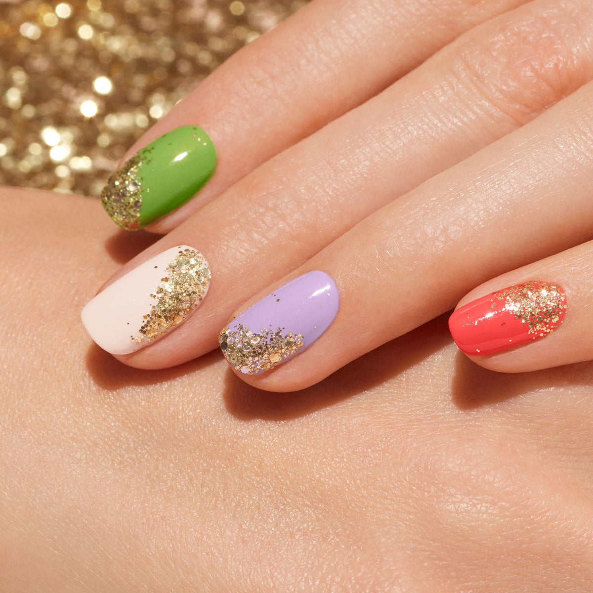 A close-up of manicured nails in green, white, lavender, and coral, each adorned with Manucurist Gold biodegradable glitter at the base. The sparkling nail art stands out against a blurred golden glitter background.