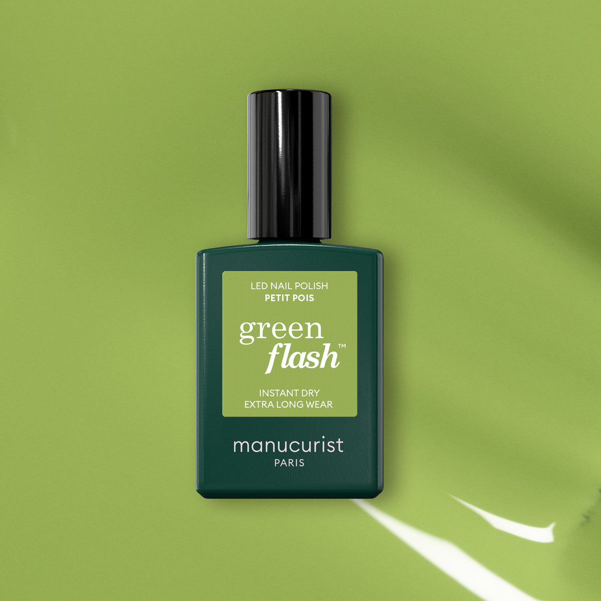 A bottle of Manucurist Green Flash gel polish in Sweet Pea rests on a green background, casting a shadow. The dark green bottle with black cap and light green label suggests bold, vibrant nails from this shade.