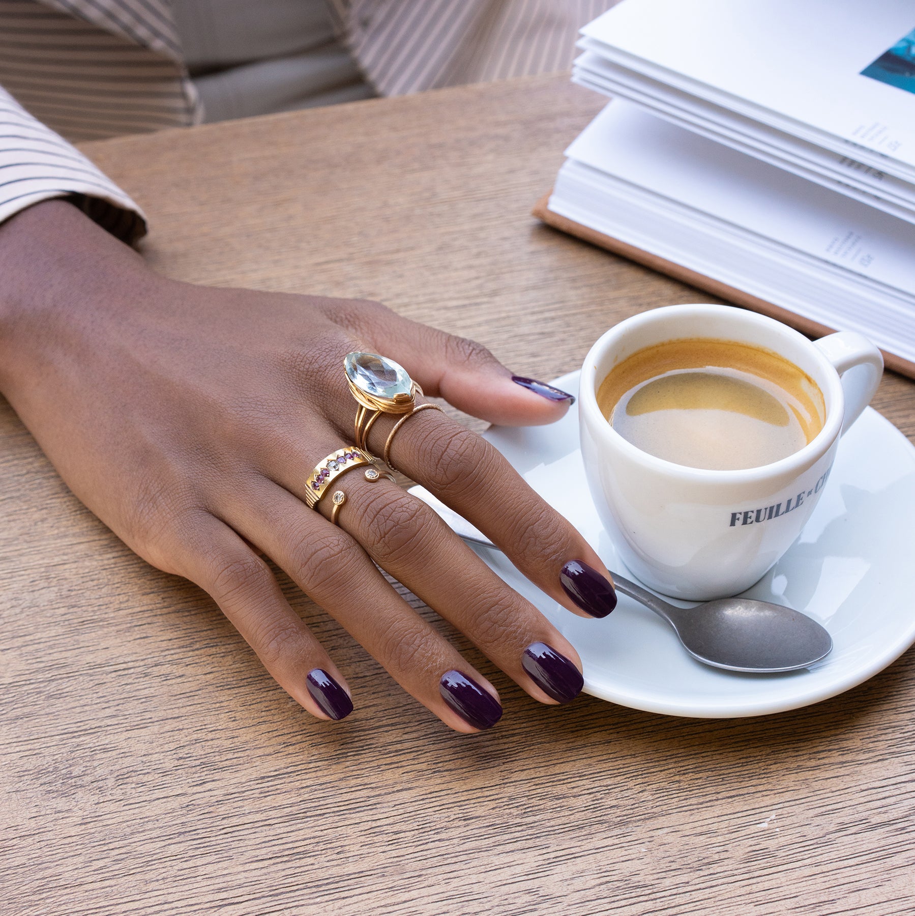 A hand with Manucurist’s Aubergine nail polish and several gold rings rests on a wooden table beside a cup of espresso, a spoon, and an open book.