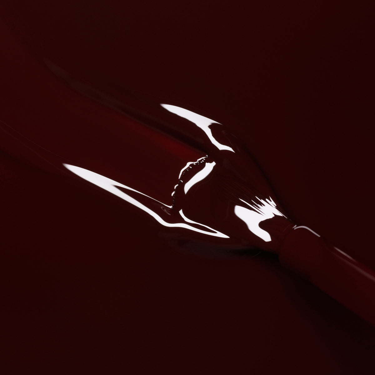 A close-up of a brush coated in Manucurist Hollyhock gel polish creates a sleek, glossy red streak across a matching surface.
