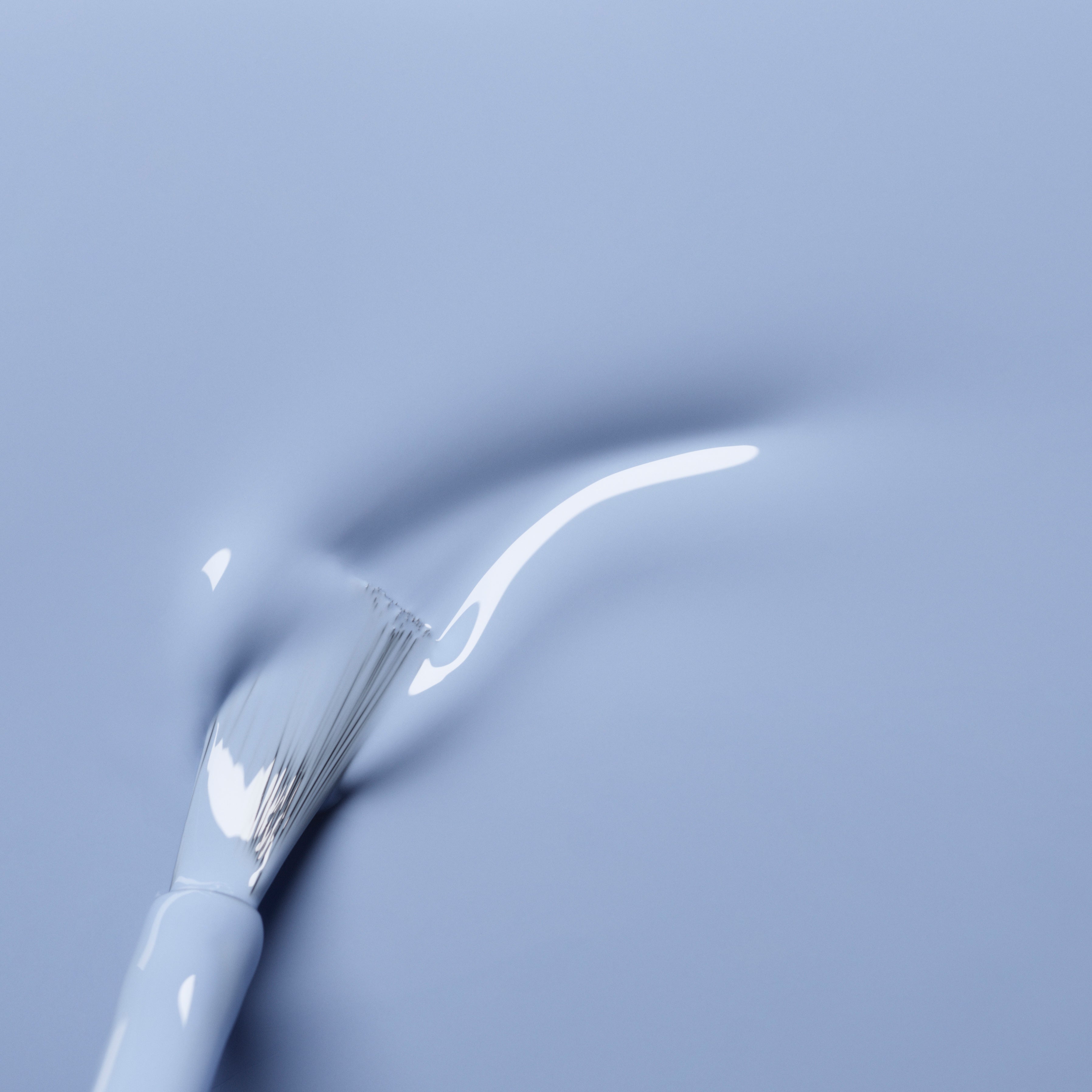 A close-up of a white nail polish brush applying glossy, pastel blue Lilas polish from Manucurist on a smooth surface. The wet, shiny finish reflects light, highlighting the beauty of this color.