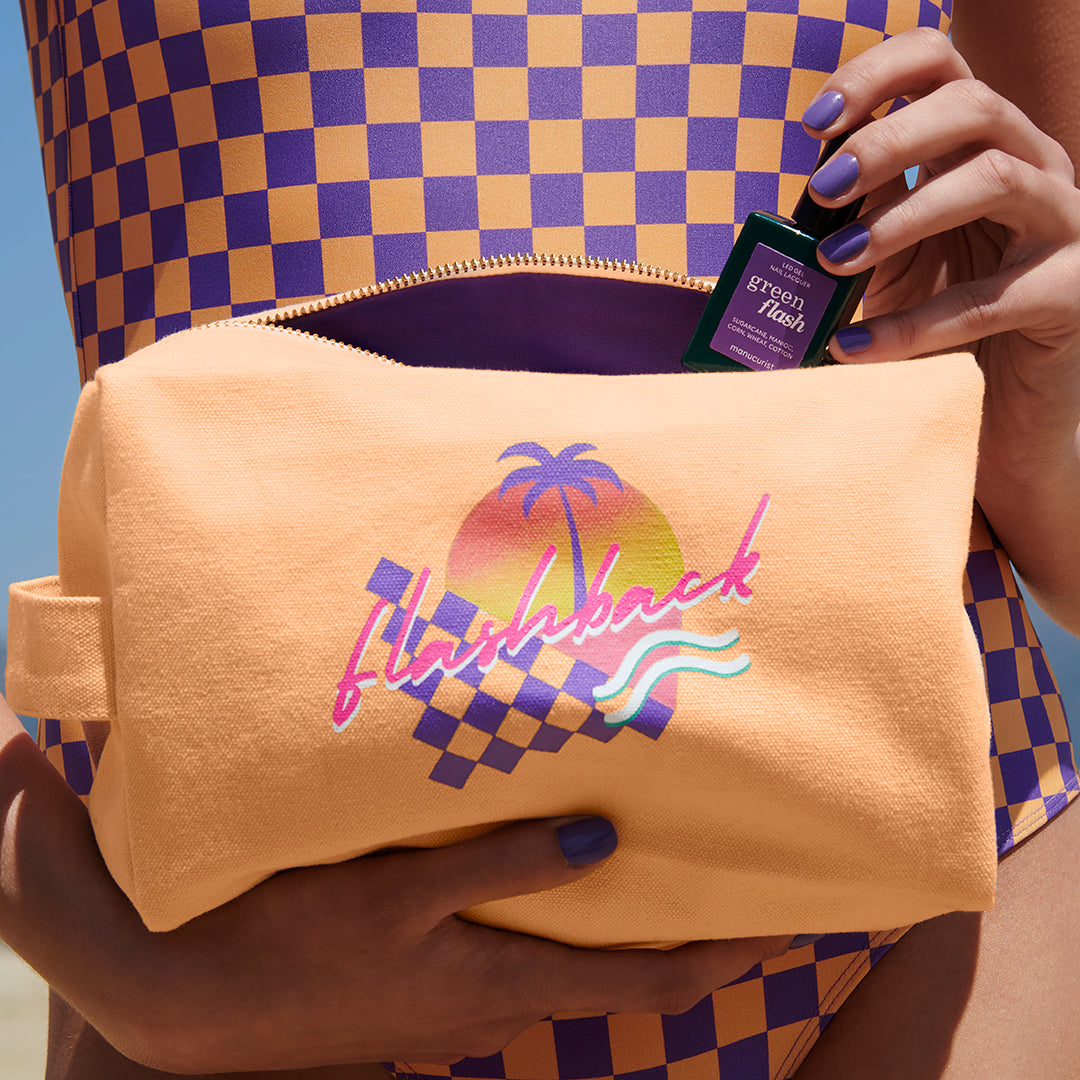 A person in a purple and yellow checkered swimsuit holds a peach bag with a palm tree and flashback design, placing a small Manucurist Green Flash gel polish bottle labeled "Iris" inside. Their nails are painted lavender.