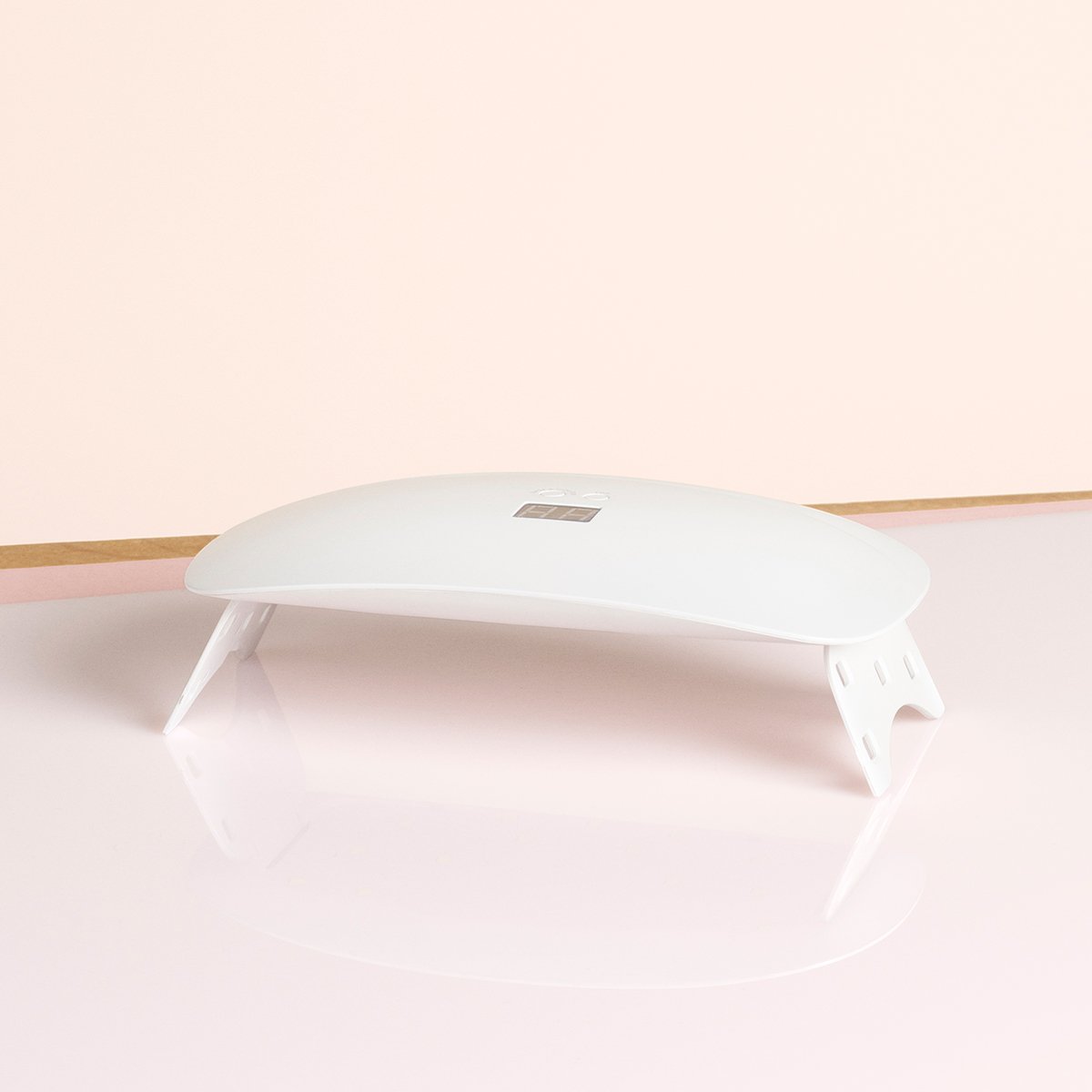The Manucurist Slim Lamp, a sleek white dome-shaped 24W LED UV manicure lamp with angled legs, rests on a glossy surface against a soft light pink background.