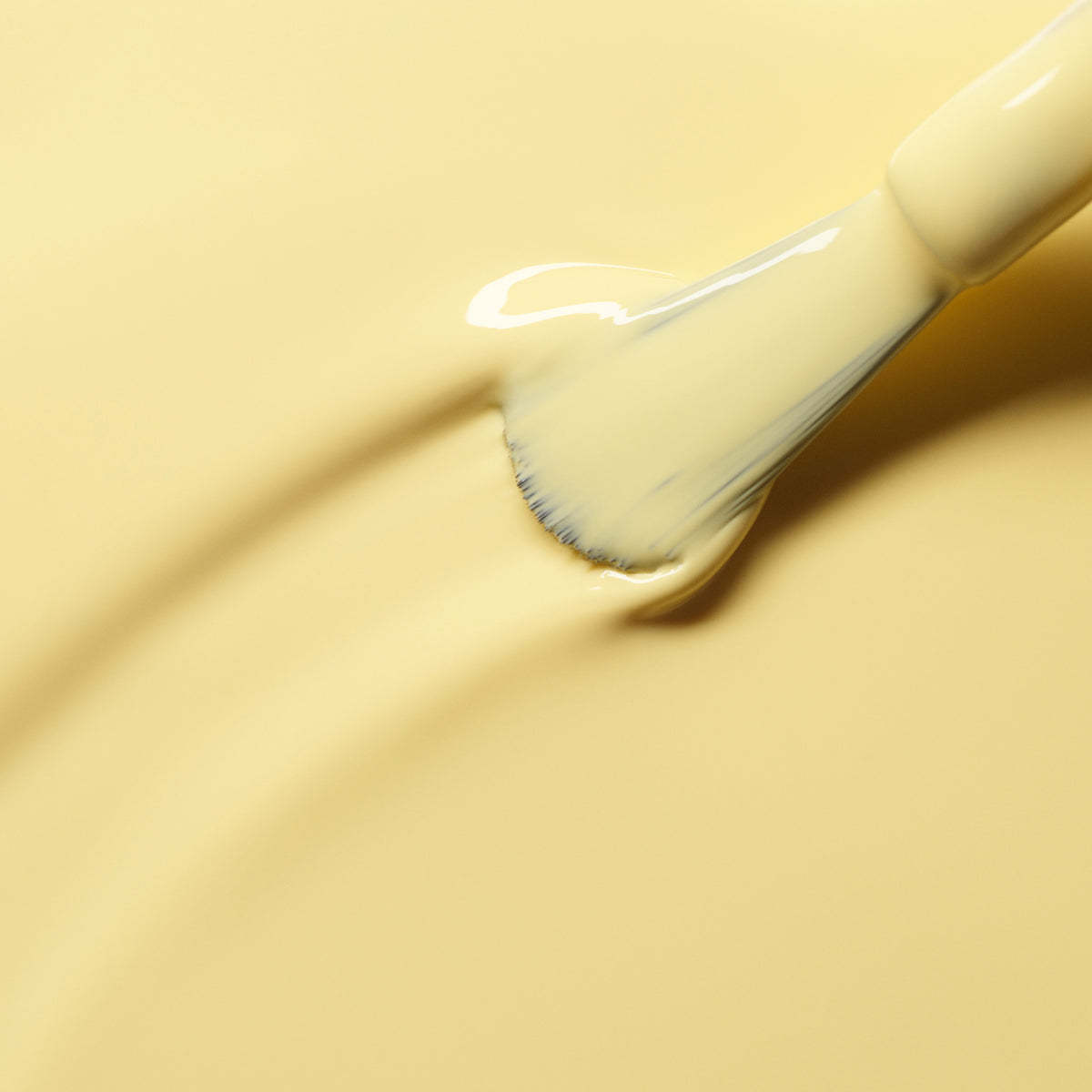 A close-up of a paintbrush spreading Manucurist Mimosa’s creamy pastel yellow paint on a flat surface, leaving soft, textured brush strokes.