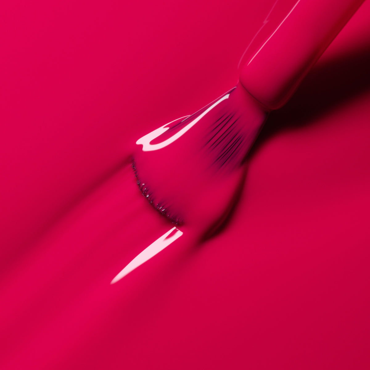 A close-up of a brush applying Manucurist Péonie gel polish, creating smooth, shiny raspberry pink strokes on a matching surface.