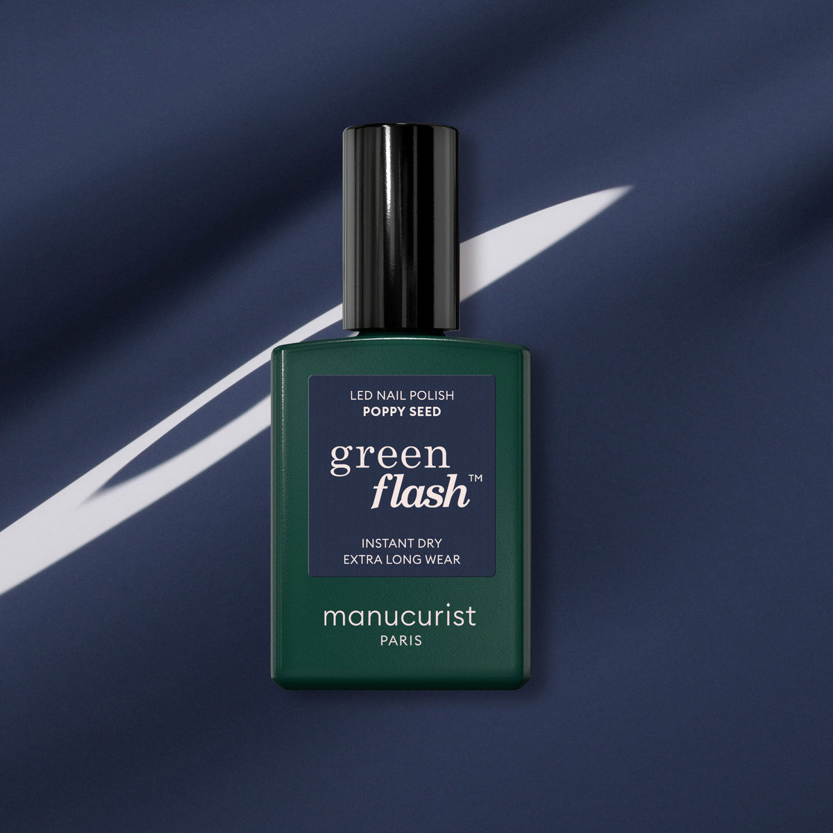 A green rectangular bottle of Manucurist Poppy Seed gel polish with a black cap sits on dark blue fabric featuring a white diagonal stripe.