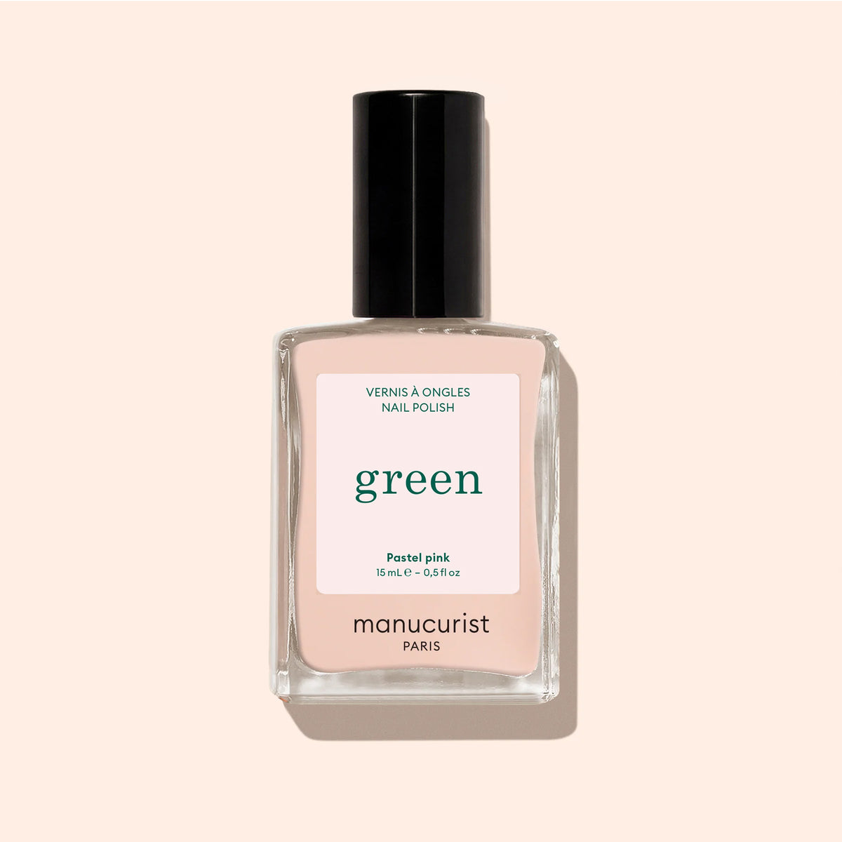 A clear glass bottle of Manucurist Pastel Pink nail polish with a black cap and beige undertones sits against a pale peach background. The label reads "Nail Polish Pastel Pink, Quick Dry Intense Color, manucurist PARIS.